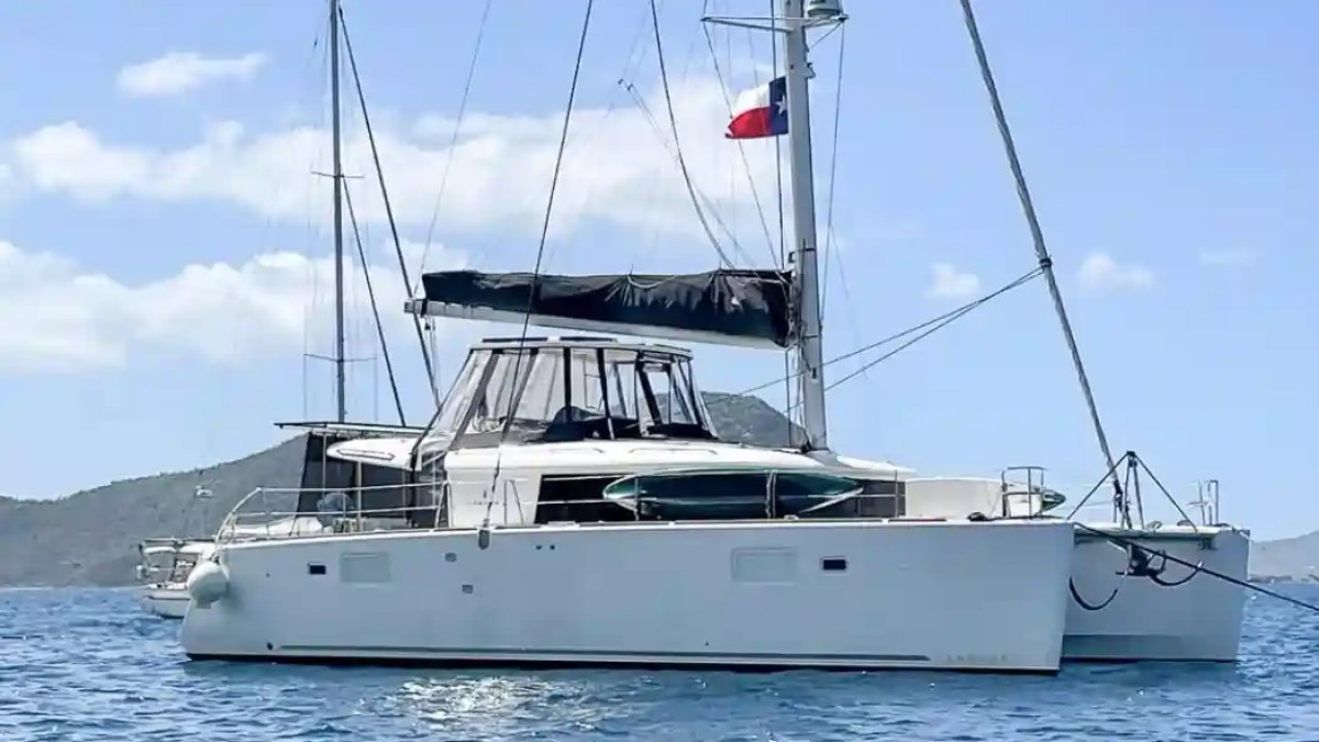 White catamaran with black sail and flag on water, mountains in background under blue sky.
