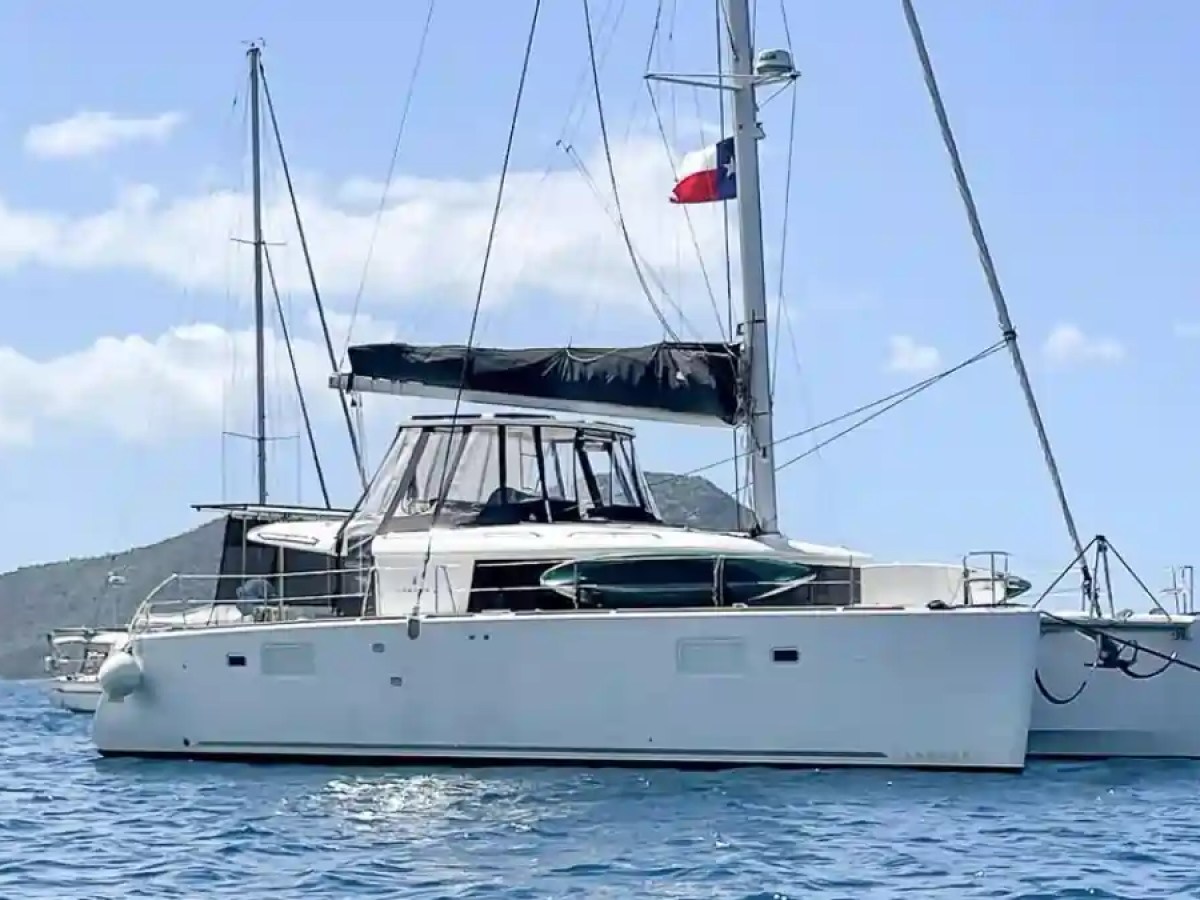 White catamaran with black sail and flag on water, mountains in background under blue sky.