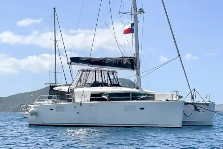 White catamaran with black sail and flag on water, mountains in background under blue sky.