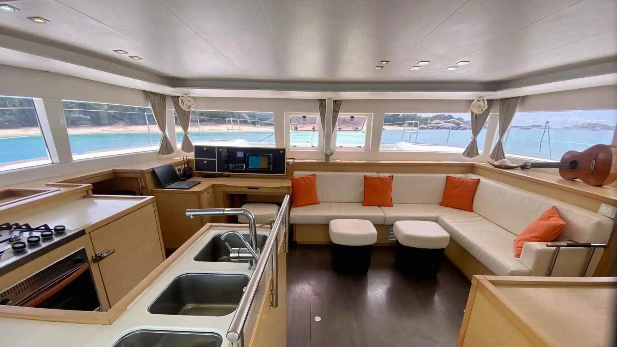 Luxury yacht interior with kitchen, lounge, and sea view through large windows, accented with orange pillows.