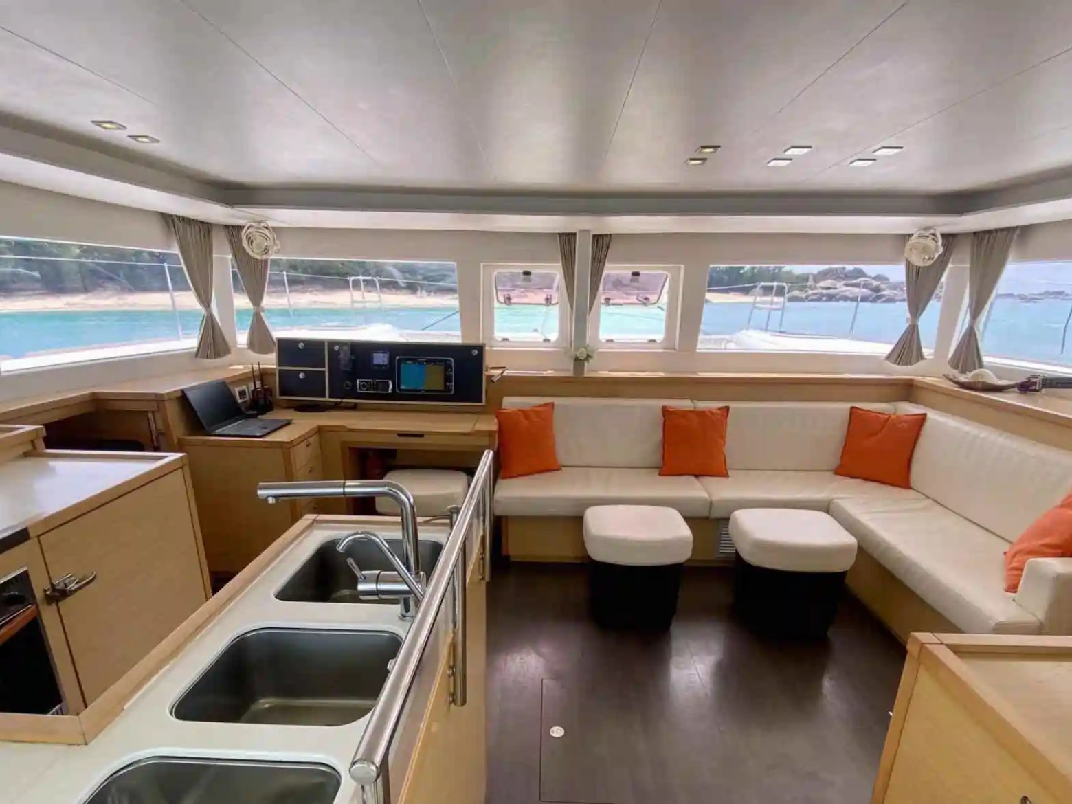 Luxury yacht interior with kitchen, lounge, and sea view through large windows, accented with orange pillows.