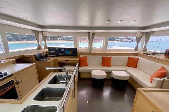 Luxury yacht interior with kitchen, lounge, and sea view through large windows, accented with orange pillows.