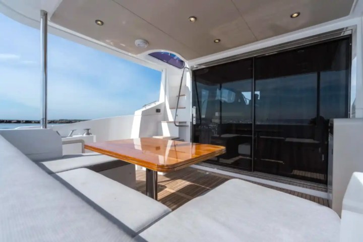 Yacht deck with a wooden table, cushioned seating, and glass doors under a blue sky.