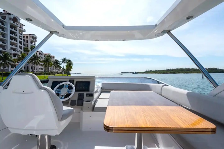 View from a yacht deck with steering wheel and table, overlooking water and buildings on a sunny day.