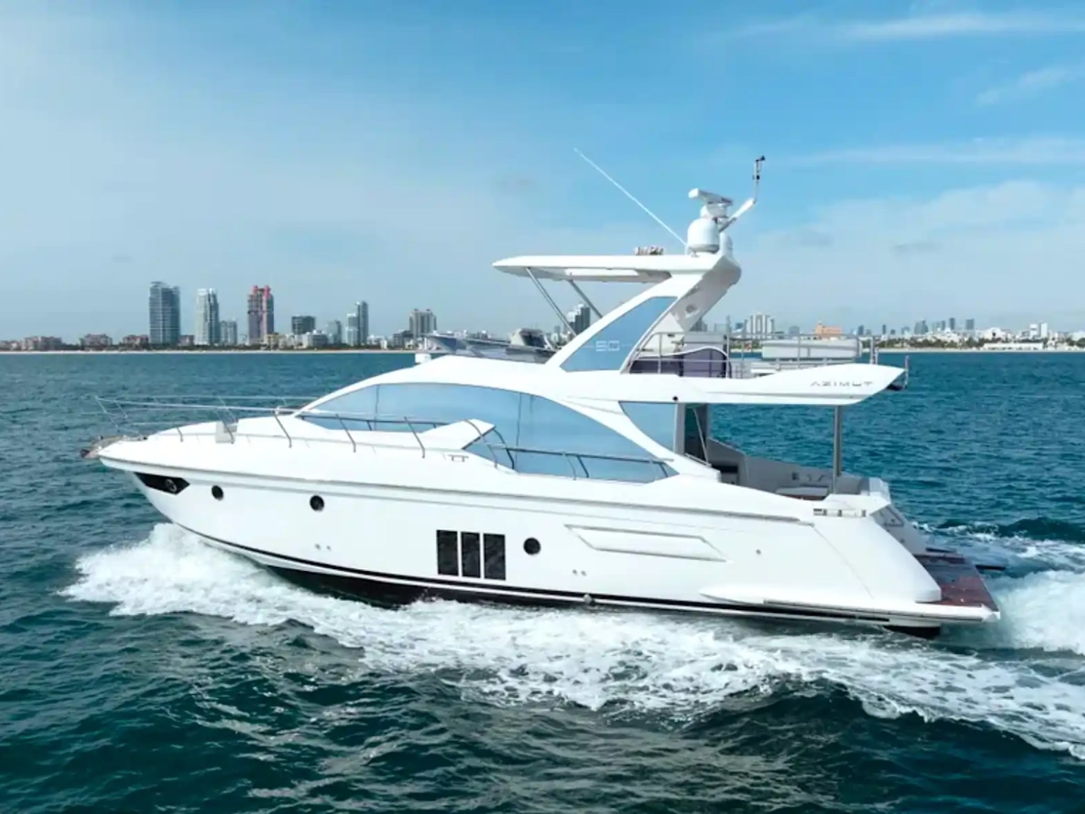 A white yacht cruising in clear blue water with a city skyline in the background.