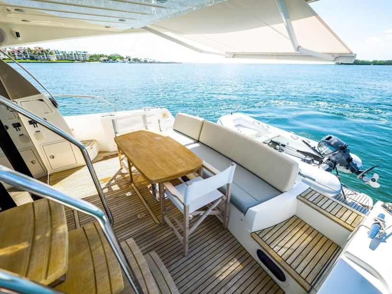 Back deck of yacht with wooden table, chairs, and view of calm sea and distant shore.
