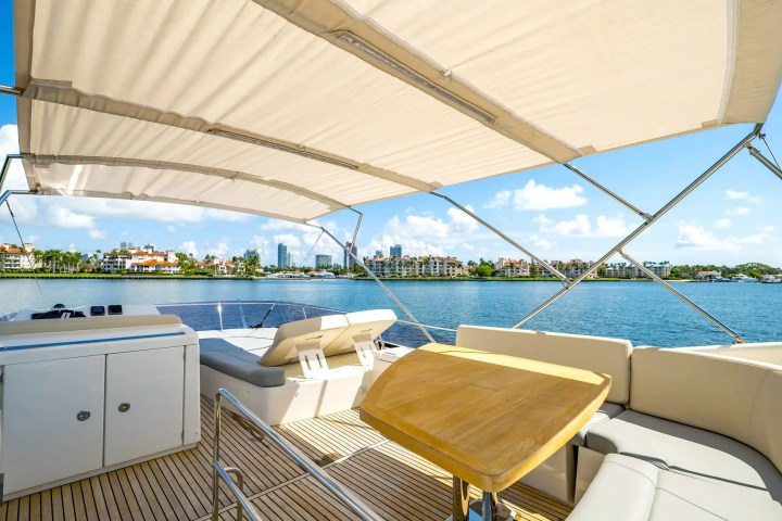 Luxury yacht deck with seating and table, under a canopy, overlooking water and distant buildings.