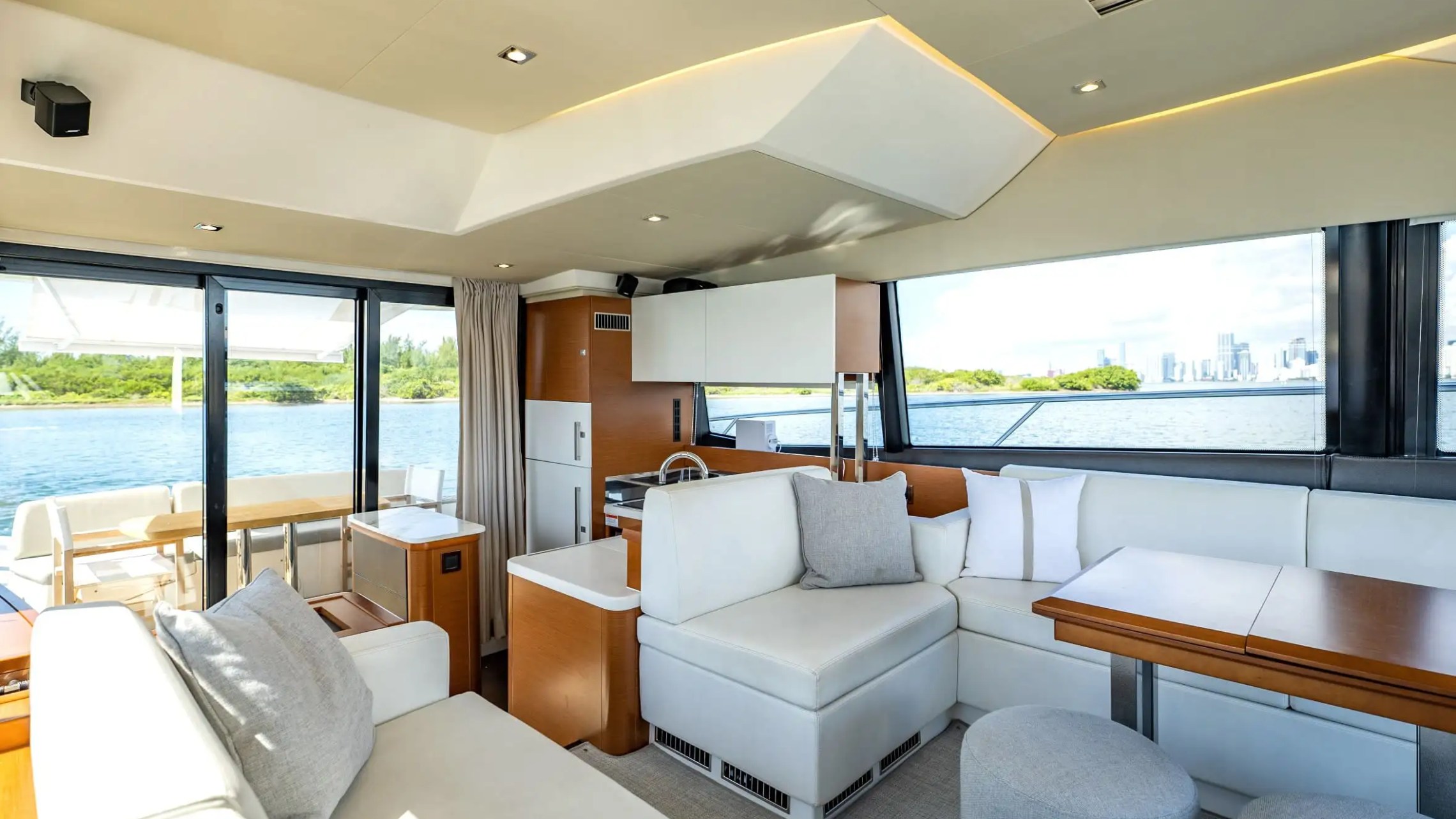 Luxury yacht interior with white seating, tables, and large windows overlooking water and greenery.