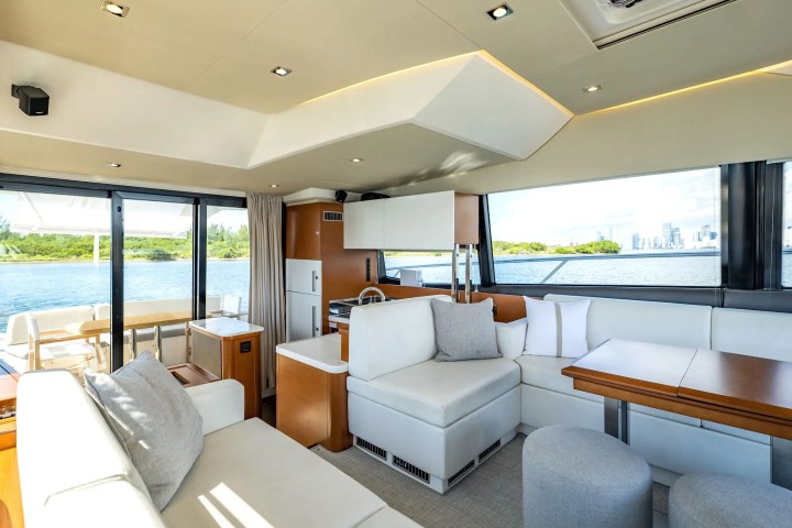 Luxury yacht interior with white seating, tables, and large windows overlooking water and greenery.