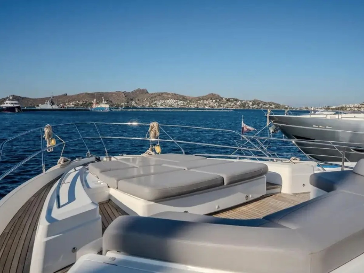 Private yacht deck with sun loungers overlooking a harbor and hills under a clear blue sky.