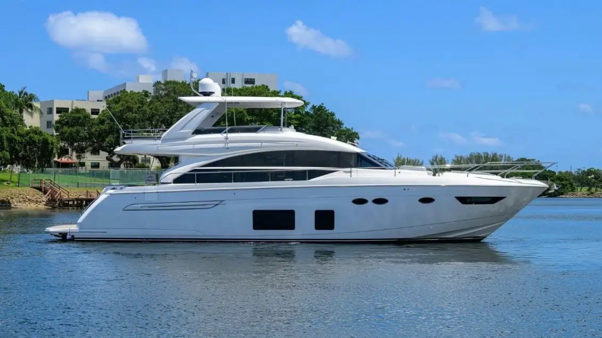 White yacht on calm water with blue sky and greenery in background.