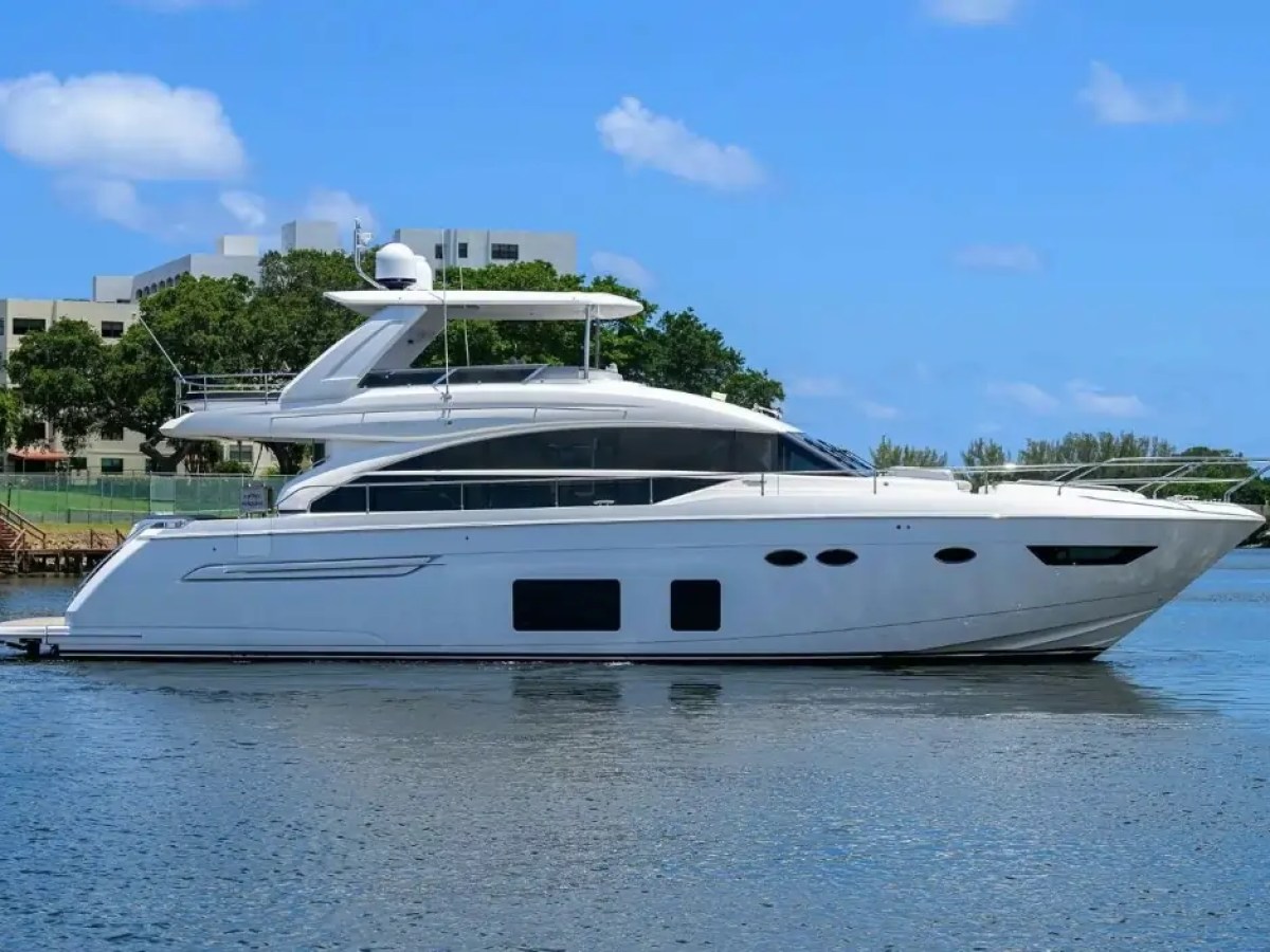 White yacht on calm water with blue sky and greenery in background.