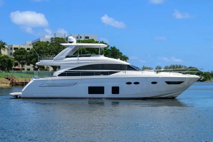White yacht on calm water with blue sky and greenery in background.