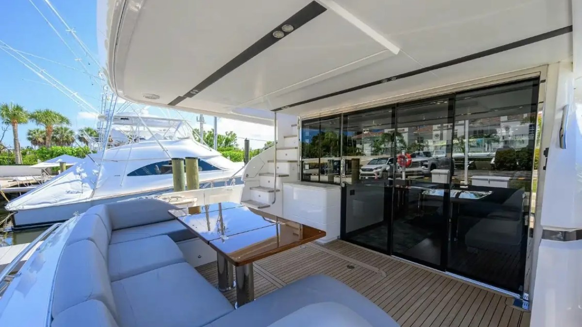 Luxury yacht deck with seating, table, and sliding glass doors to interior, docked near palm trees.
