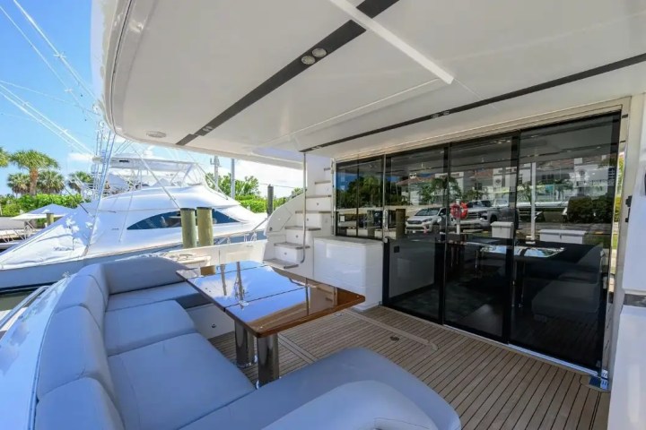 Luxury yacht deck with seating, table, and sliding glass doors to interior, docked near palm trees.