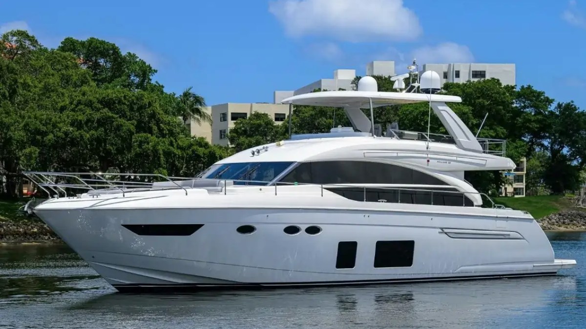 Luxurious white yacht sailing on calm water, trees and buildings in the background.