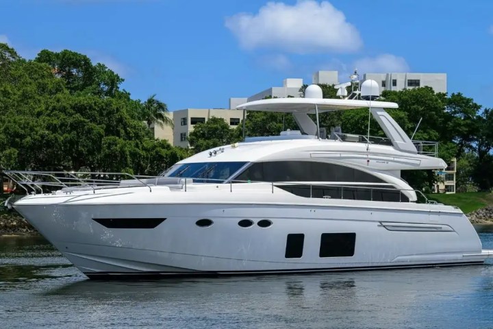 Luxurious white yacht sailing on calm water, trees and buildings in the background.