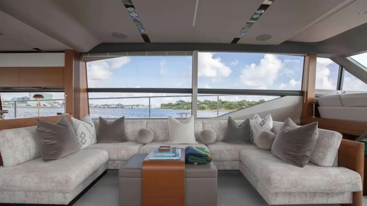 Luxurious yacht interior with beige sofas, cushions, and large windows overlooking water and blue sky.