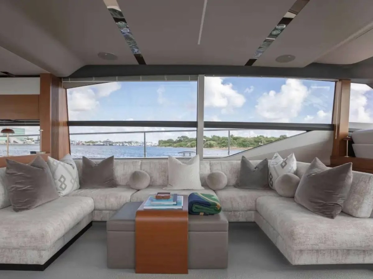 Luxurious yacht interior with beige sofas, cushions, and large windows overlooking water and blue sky.