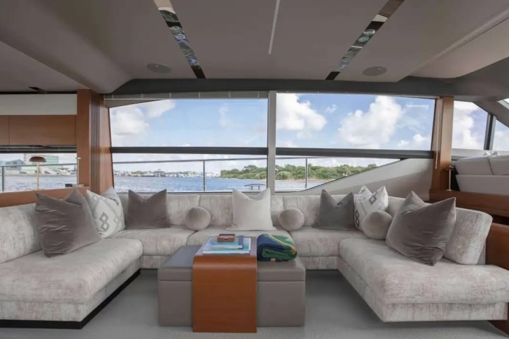 Luxurious yacht interior with beige sofas, cushions, and large windows overlooking water and blue sky.
