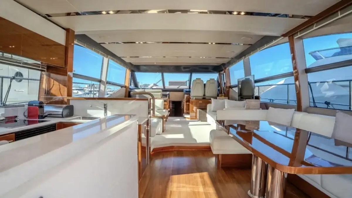 Luxurious yacht interior with white seating, wooden floors, and large windows showing a marina view.