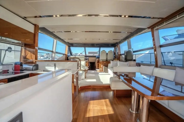 Luxurious yacht interior with white seating, wooden floors, and large windows showing a marina view.