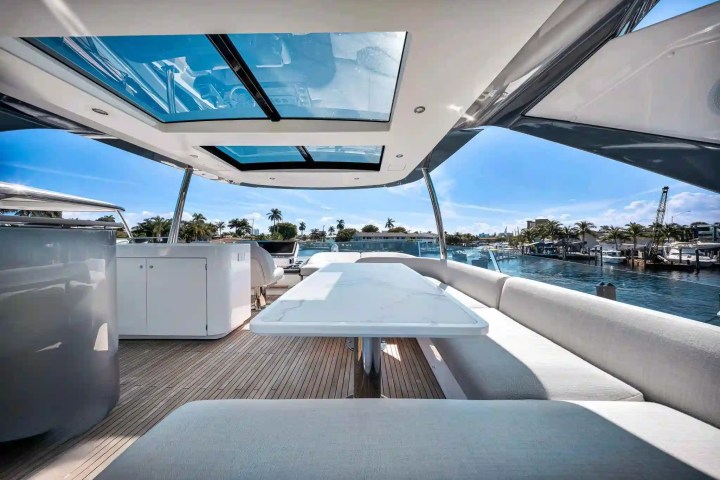 Luxury yacht deck with white seating and table under skylights, view of palm trees and water.