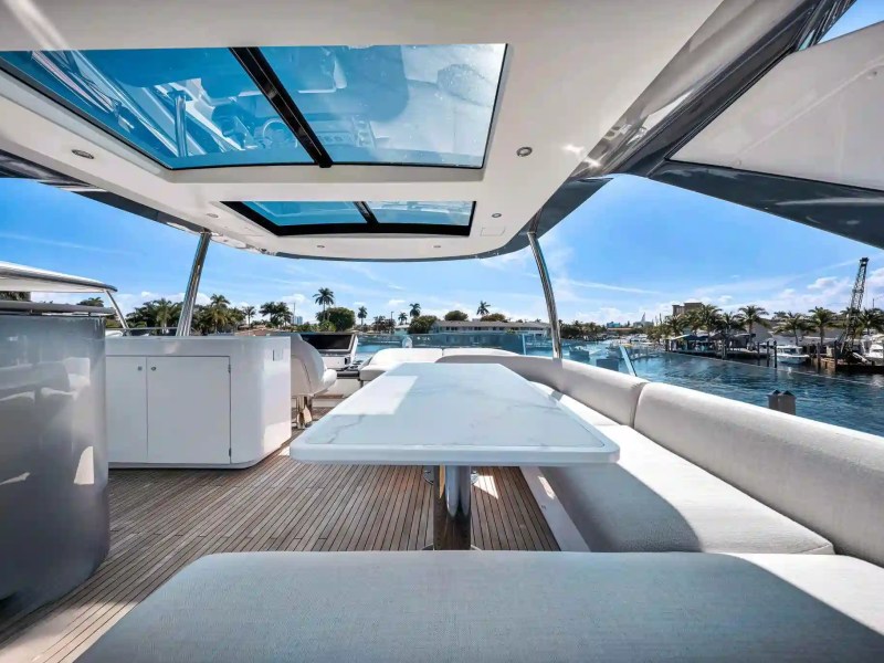Luxury yacht deck with white seating and table under skylights, view of palm trees and water.