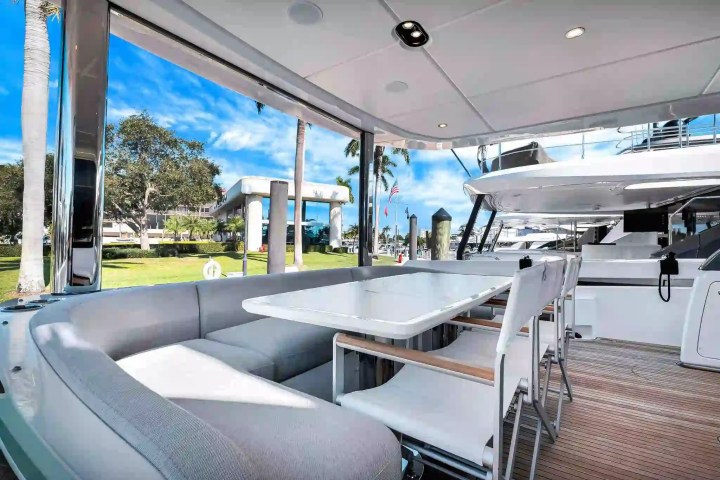 Outdoor yacht dining area with table, chairs, and sofa, overlooking dock and palm trees against a clear sky.
