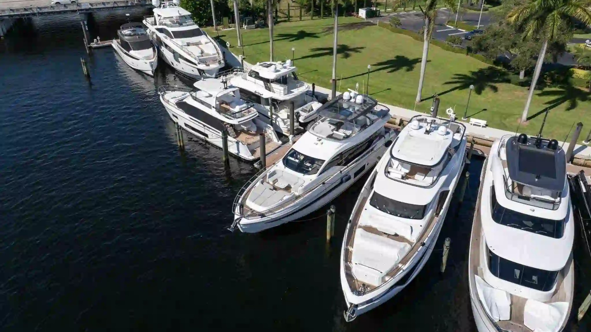 Aerial view of several white yachts docked next to a grassy area lined with palm trees.