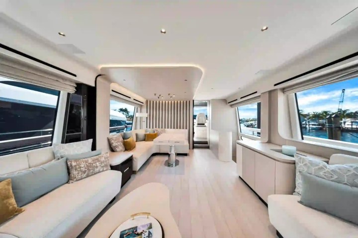 Luxurious boat interior with white seating, large windows, and a view of a marina outside.
