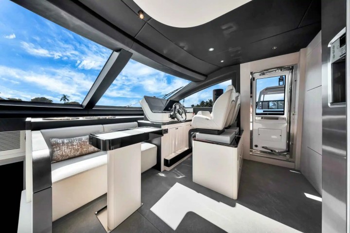 Modern yacht interior with helm, white seating, table, and large windows showing blue sky and palm trees.