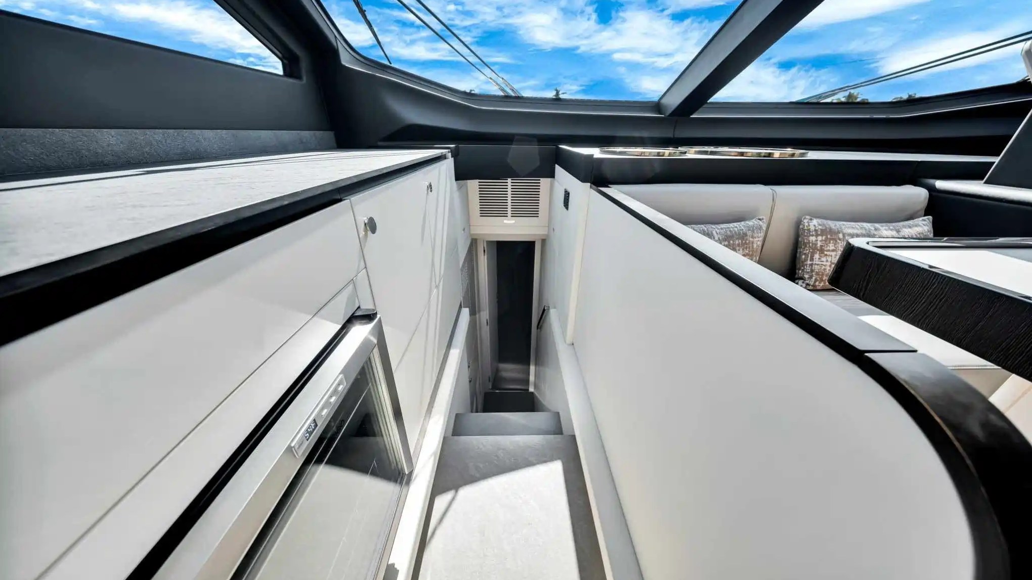 Interior of a modern boat with sleek white cabinetry, a small fridge, and large window with view of blue sky.