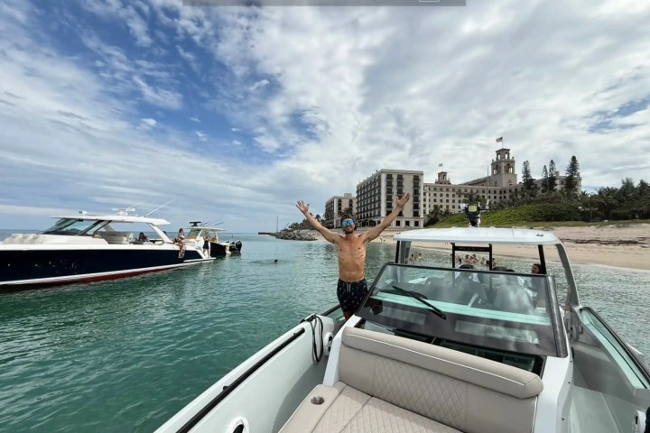 Person on a boat with arms raised, near a beach and a large hotel in the background.