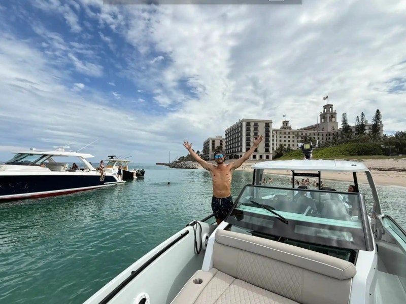 Person on a boat with arms raised, near a beach and a large hotel in the background.