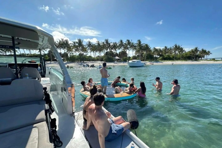 Group of people relaxing on a float and boat near tropical beach with palm trees.