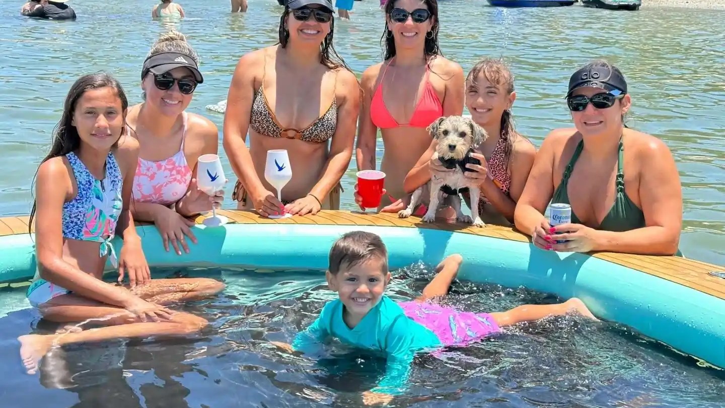Group of women and a child in a floating pool on a lake, with a dog, drinks, and beach background.