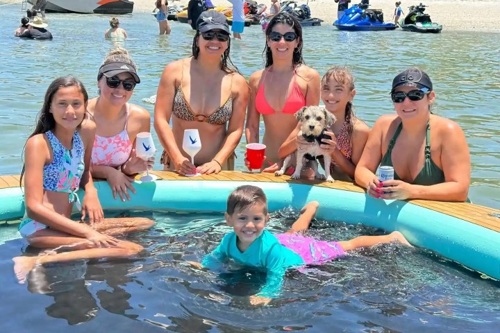 Group of women and a child in a floating pool on a lake, with a dog, drinks, and beach background.
