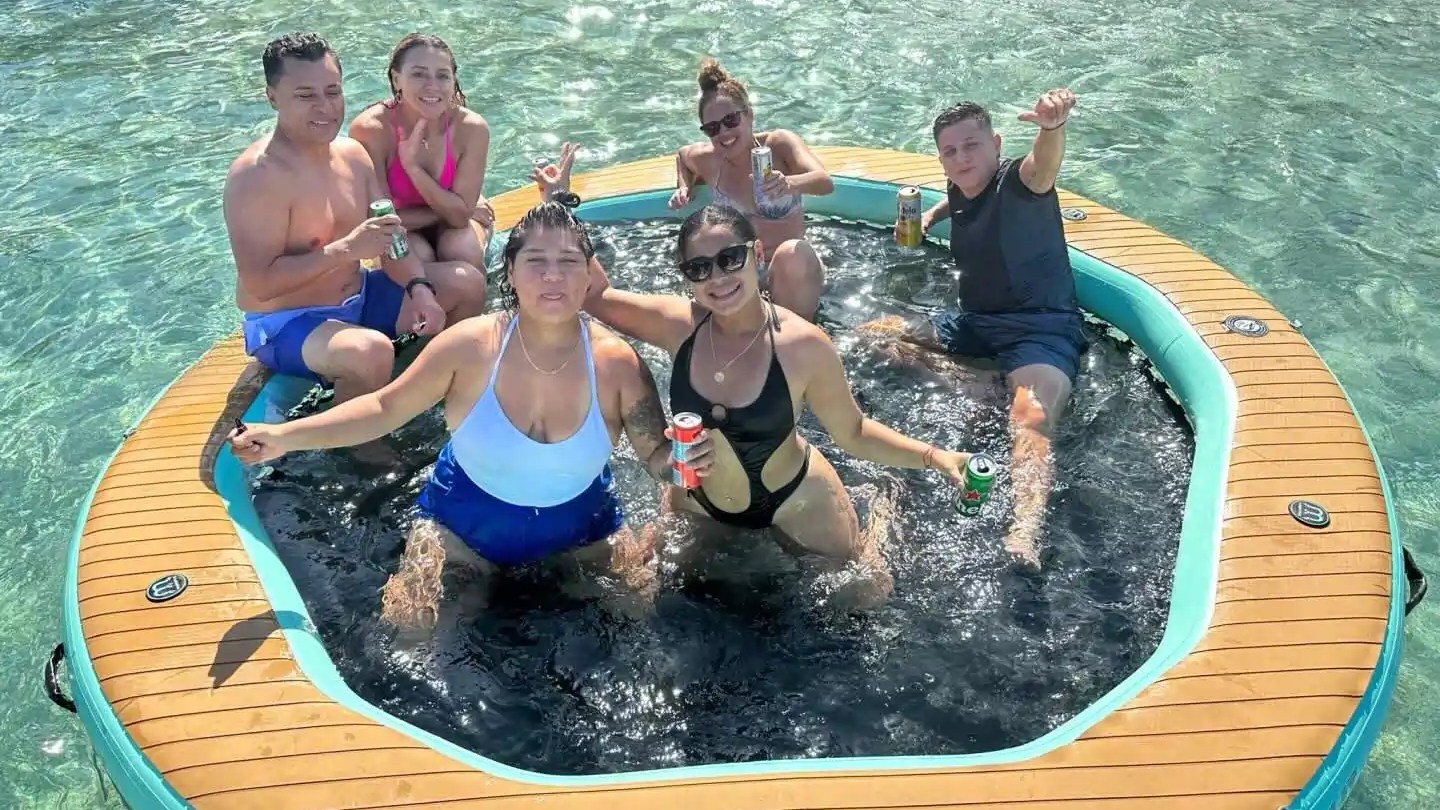 Six people on an inflatable raft in clear water, holding drinks and smiling at the camera.