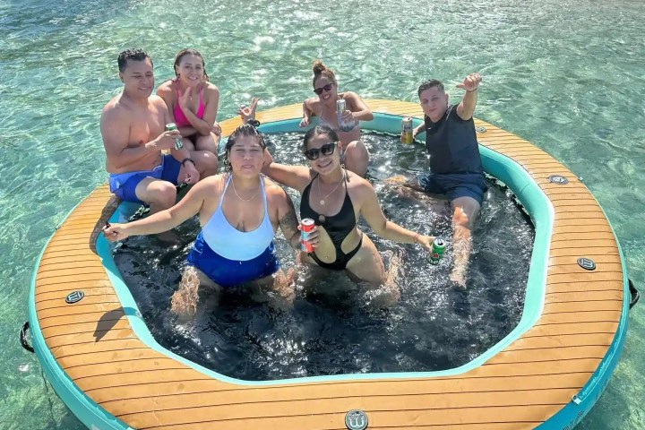 Six people on an inflatable raft in clear water, holding drinks and smiling at the camera.
