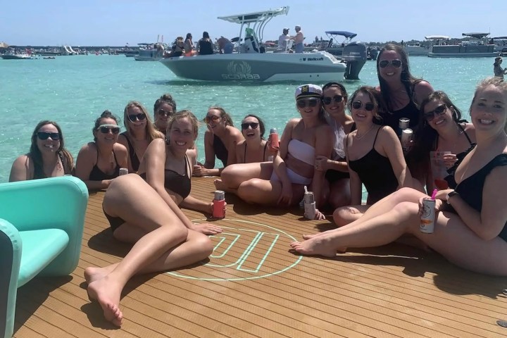 Group of people on a boat deck in swimwear, with boat and water in background.