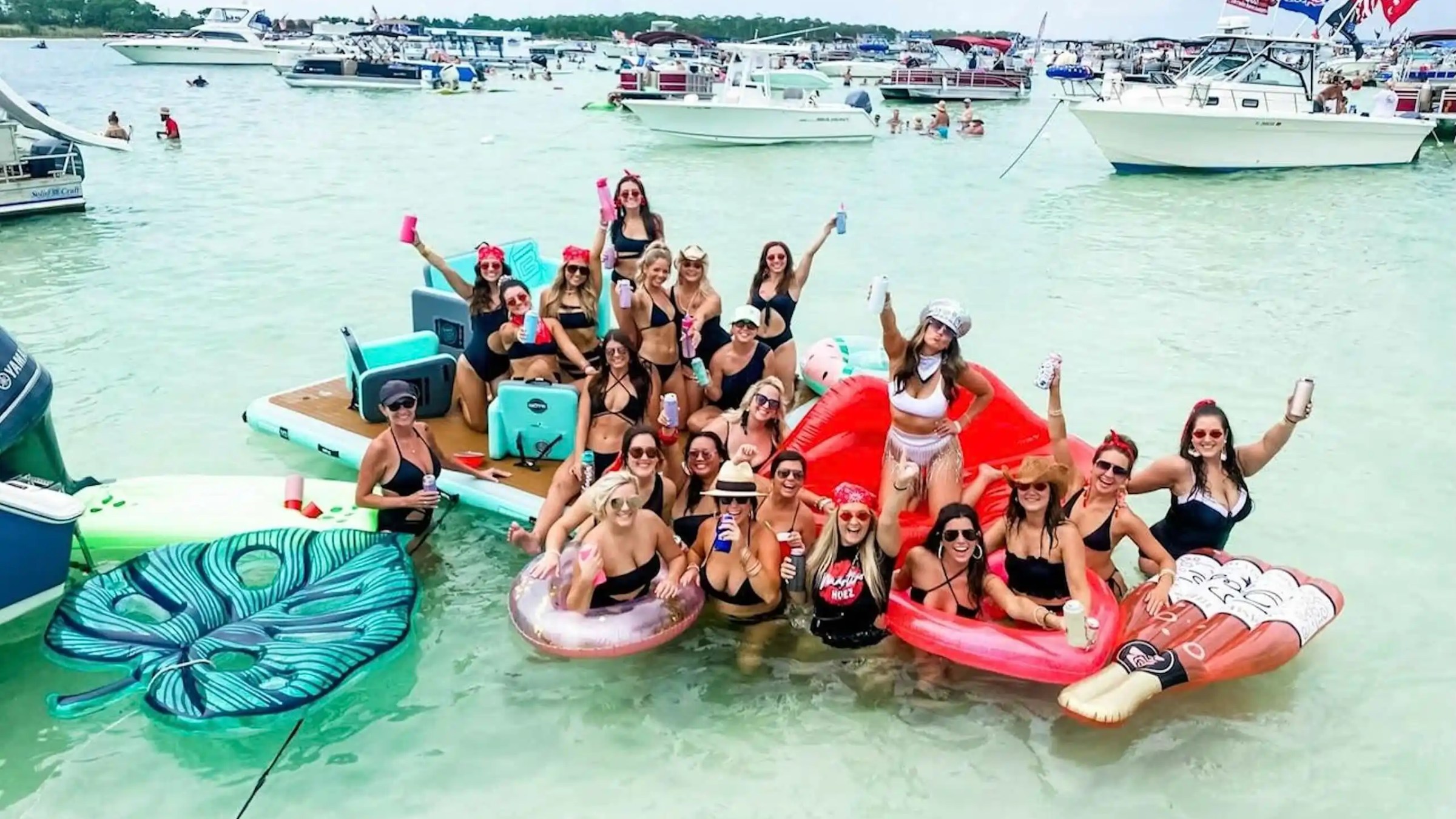 Group of women in bikinis celebrating on inflatable floats in water, boats in the background.