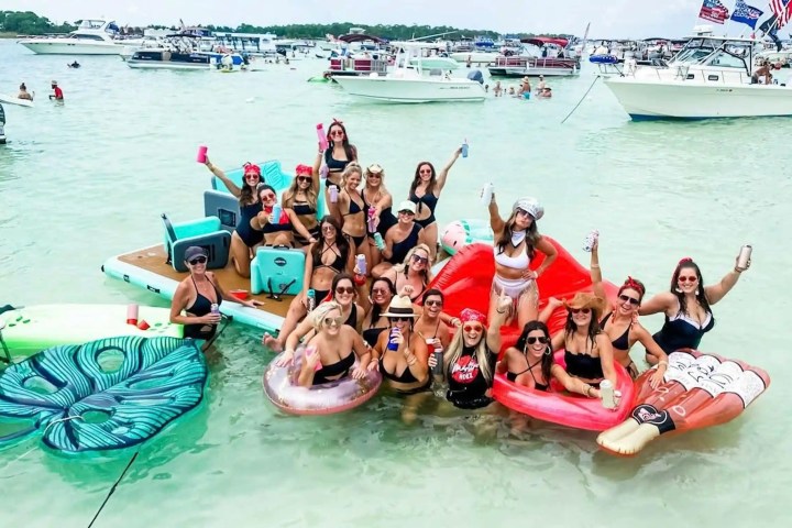 Group of women in bikinis celebrating on inflatable floats in water, boats in the background.