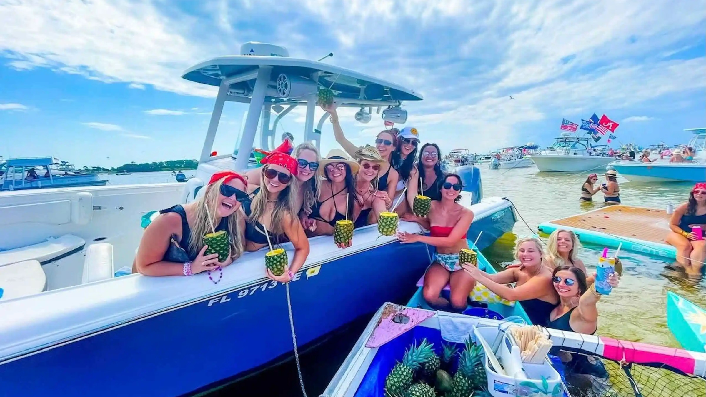 Group of women on a boat holding pineapple drinks, surrounded by water and other boats.