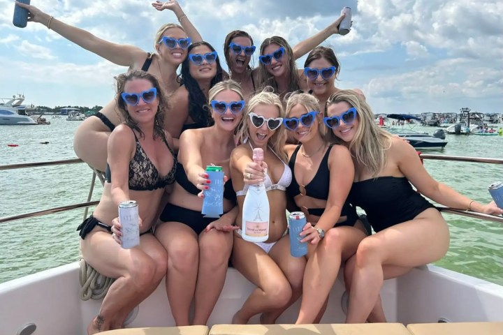 Group of women in swimsuits with heart-shaped sunglasses posing on a boat, holding drinks.