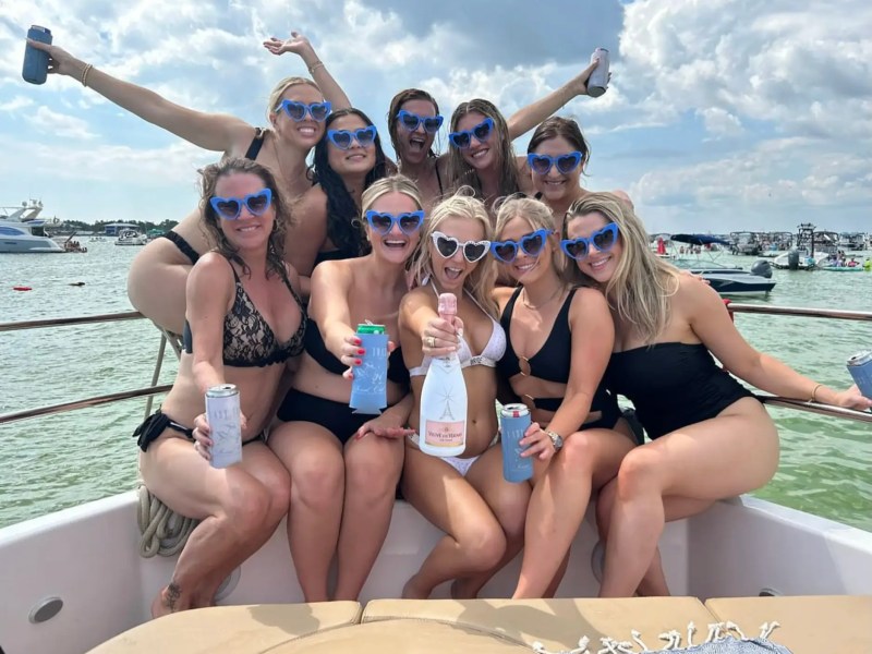 Group of women in swimsuits with heart-shaped sunglasses posing on a boat, holding drinks.