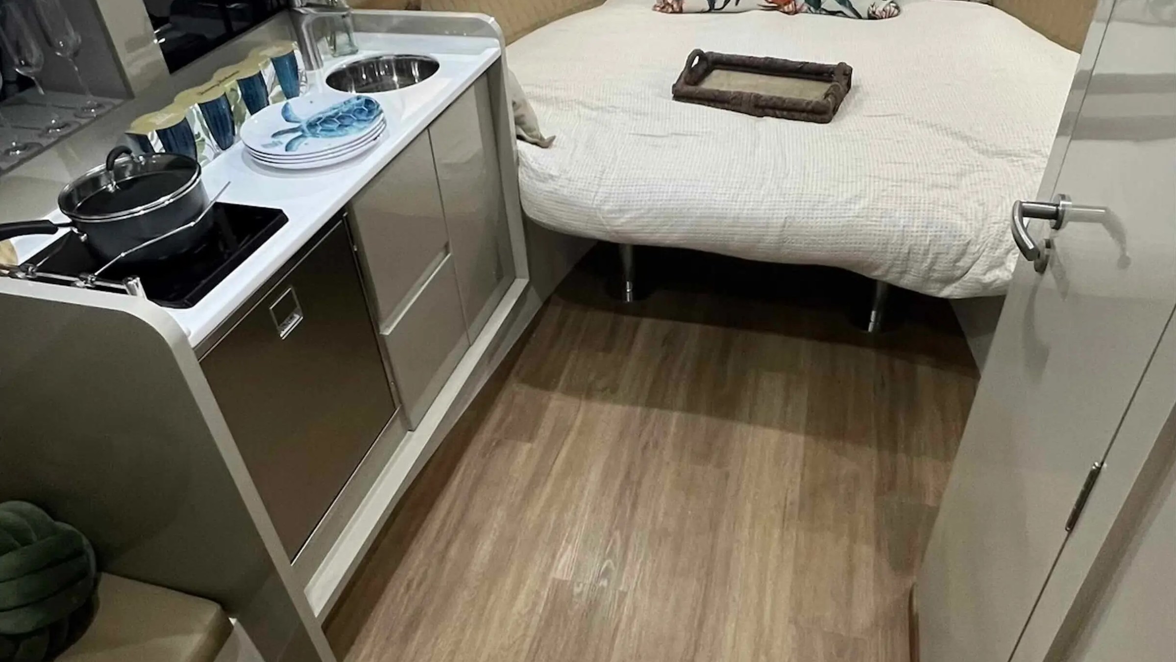 Compact boat interior with a kitchen and bed, featuring nautical-themed pillows and cookware on the stove.