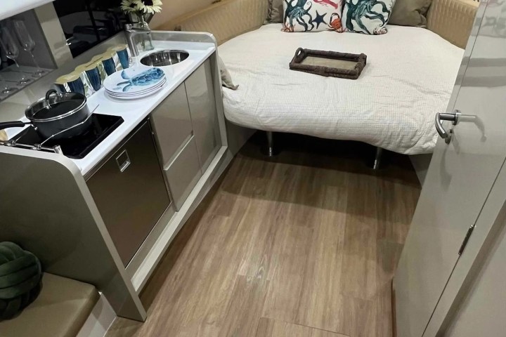 Compact boat interior with a kitchen and bed, featuring nautical-themed pillows and cookware on the stove.