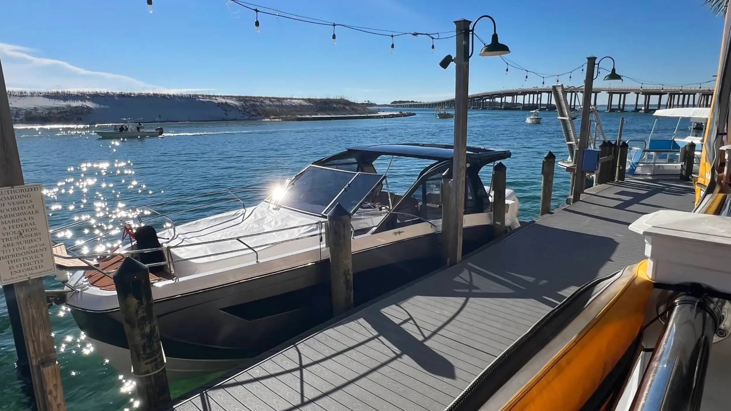 Motorboat docked at a sunny marina with string lights and a bridge in the background.