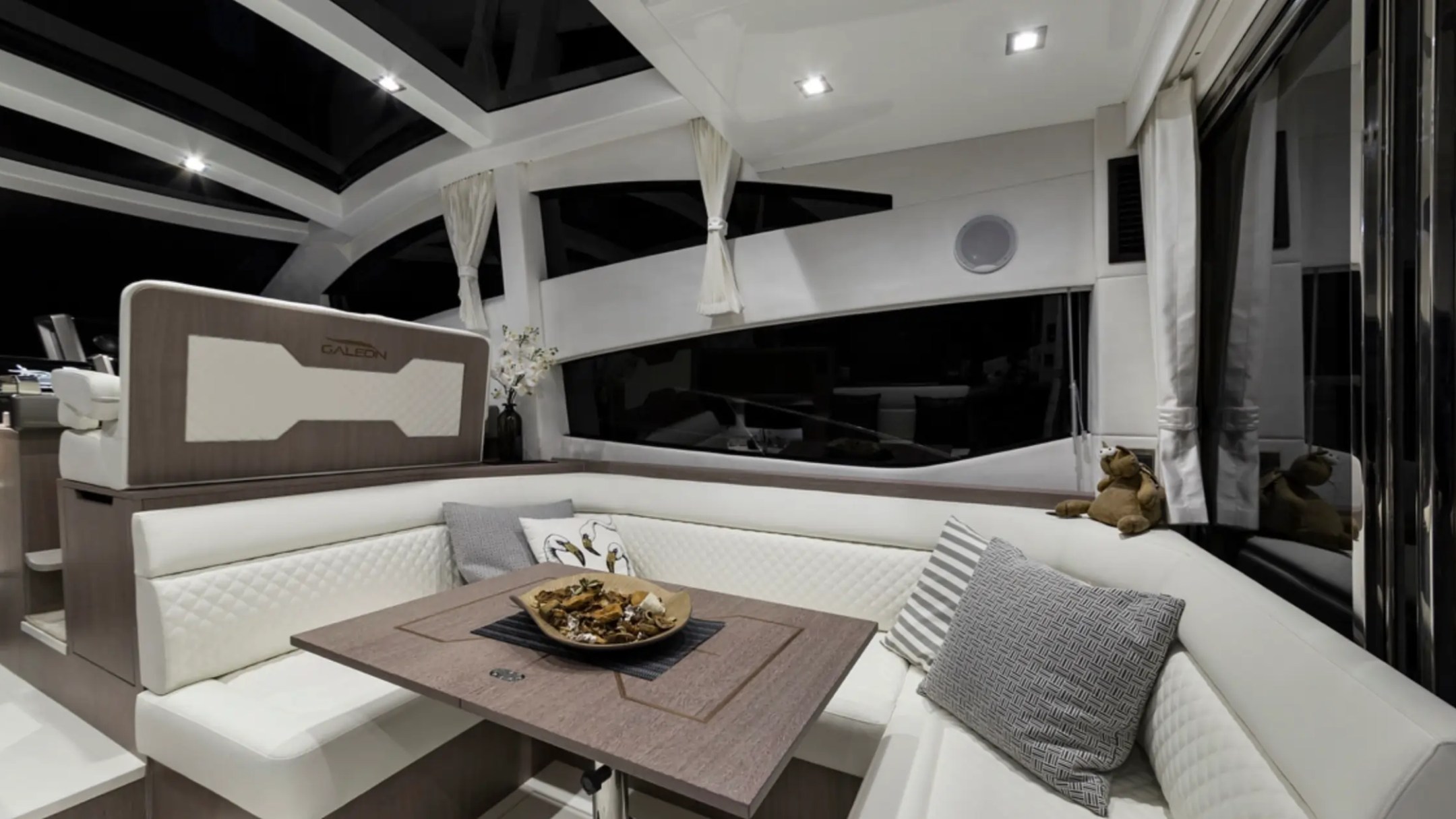 Elegant yacht interior with white seating, wooden table, and decorative pillows.